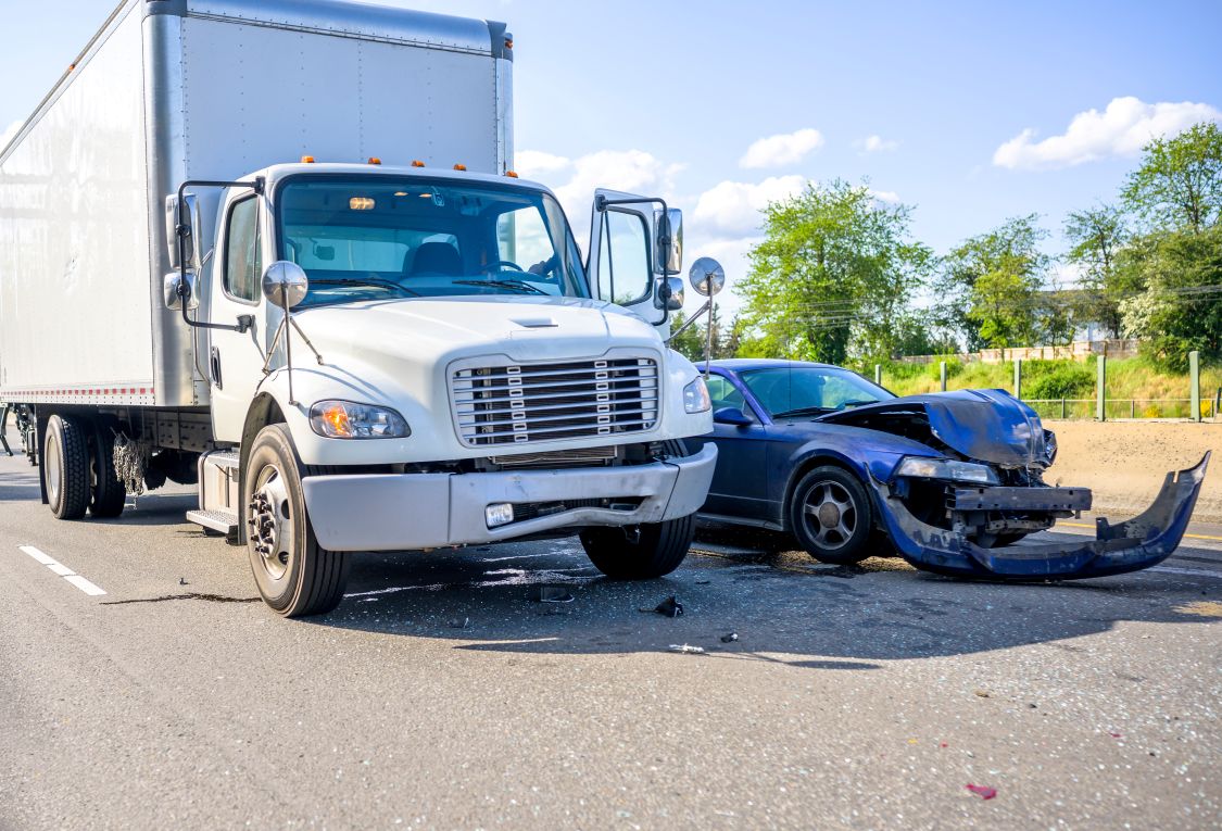 A white box truck and a heavily damaged blue sedan involved in a serious accident on a highway, with shattered glass and debris scattered on the road. The front bumper of the sedan is completely detached, illustrating the severe impact. What to Know About Box Truck Accidents: This image highlights the dangers of collisions between large commercial trucks and passenger vehicles.